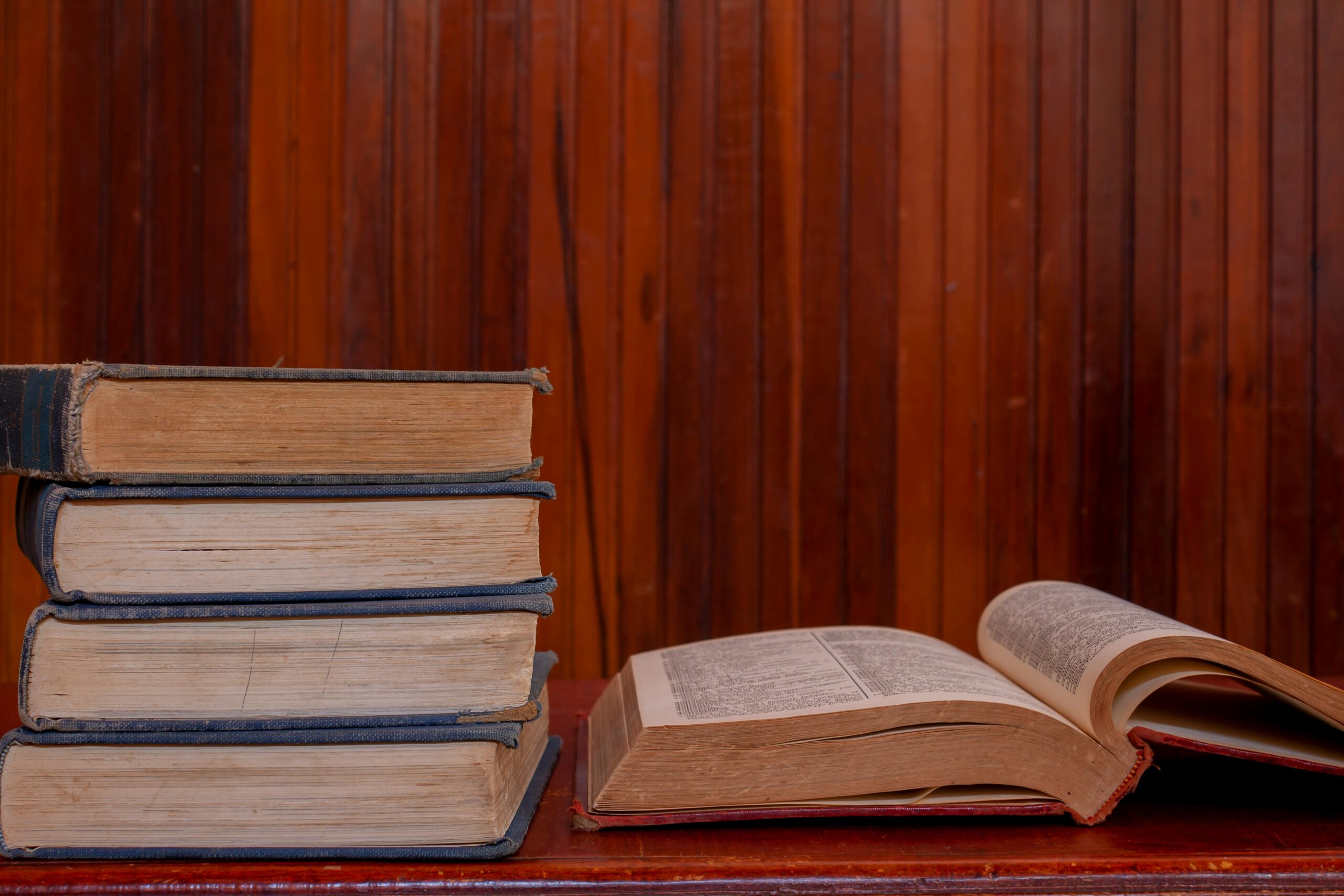 A stack of vintage books with one open, placed on a wooden table, against a wood-paneled wall.
